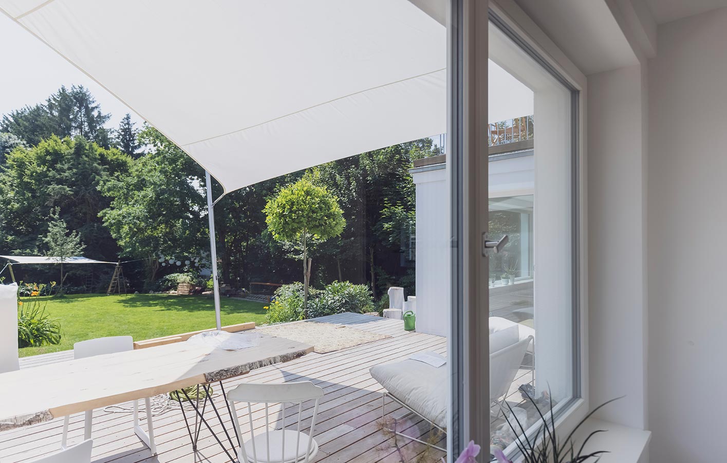 Blick aus dem Fenster in den sonnigen Garten - die Terrasse wird geschützt durch ein Sonnensegel